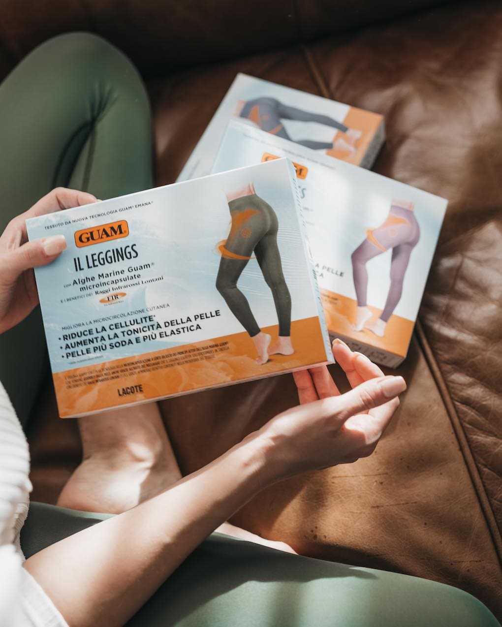 Woman in GUAM anti-cellulite leggings holding two boxes of compression leggings for cellulite on a sofa